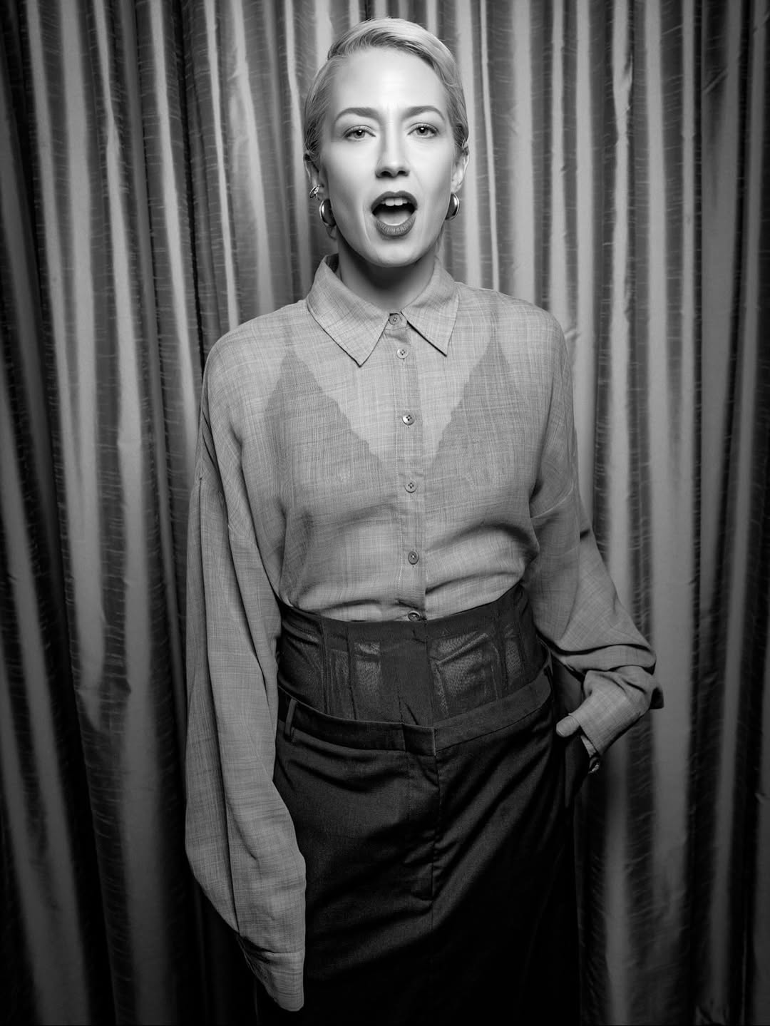 Person wearing a patterned shirt over a plain top with a wide belt, standing against a striped curtain.